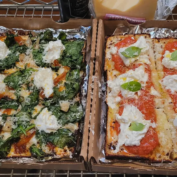 Two rectangular pan pizzas in boxes, one with greens and burrata, one with tomato, burrata and basil