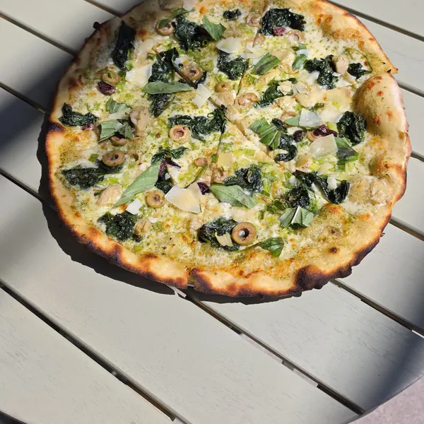A white pizza with greens, olives, artichokes, basil and shaved cheese on a patio table