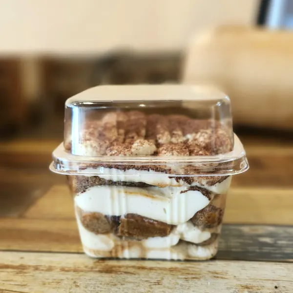 A layered tiramisu in a clear takeout cup, dusted with cocoa