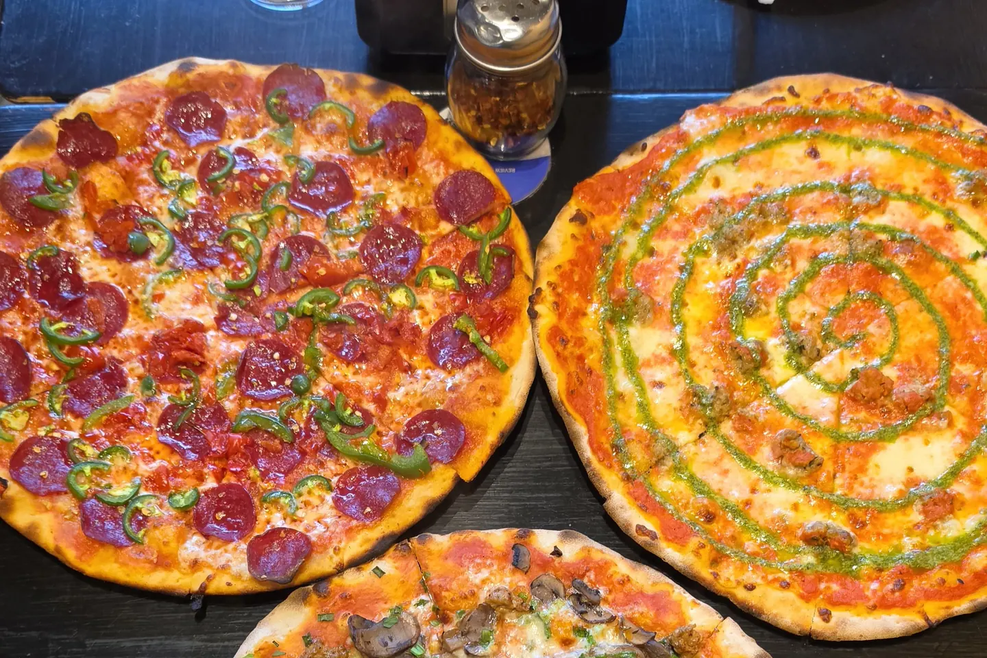 Three thin-crust pizzas on a dark table: the Diva on the left, the Jesse P. with beef meatball and a pesto swirl on the right, and a mushroom pie in front